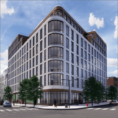 A large, modern multi-story building with white exterior and numerous large windows on a street corner. The building has trees and people walking around its ground level. The sky above is clear with a few clouds.