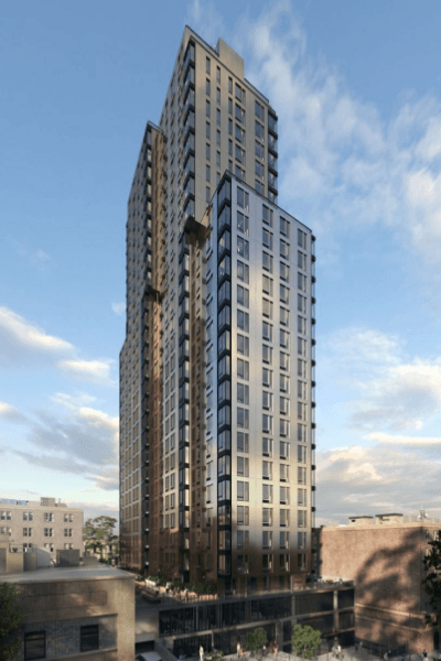 Tall, modern skyscraper with reflective glass windows and multiple sections, towering over surrounding low-rise buildings against a partly cloudy blue sky. Trees and small buildings are visible at the base.