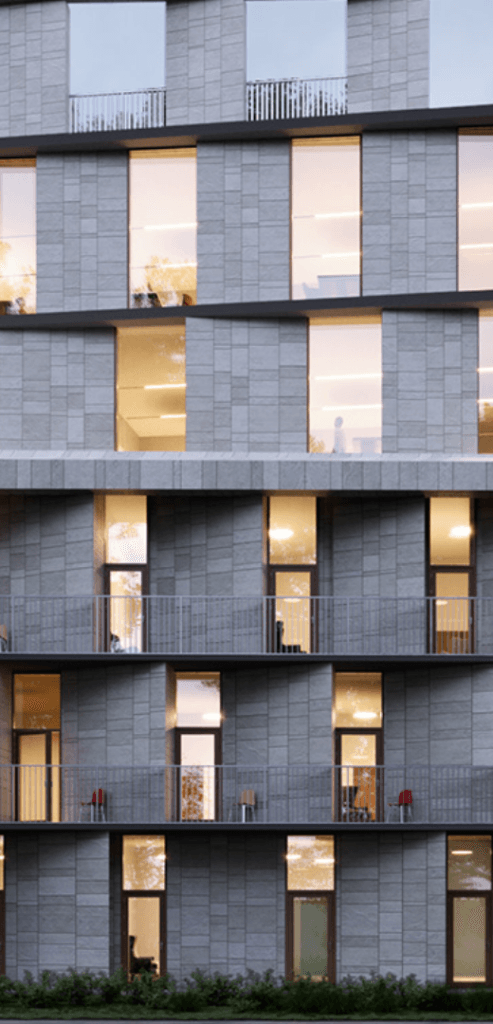 A modern multi-story building with a facade of rectangular patterns and large windows. The building has a mix of illuminated and dark windows. The lower floors feature balconies with metal railings. Sparse vegetation is visible at the base of the building.