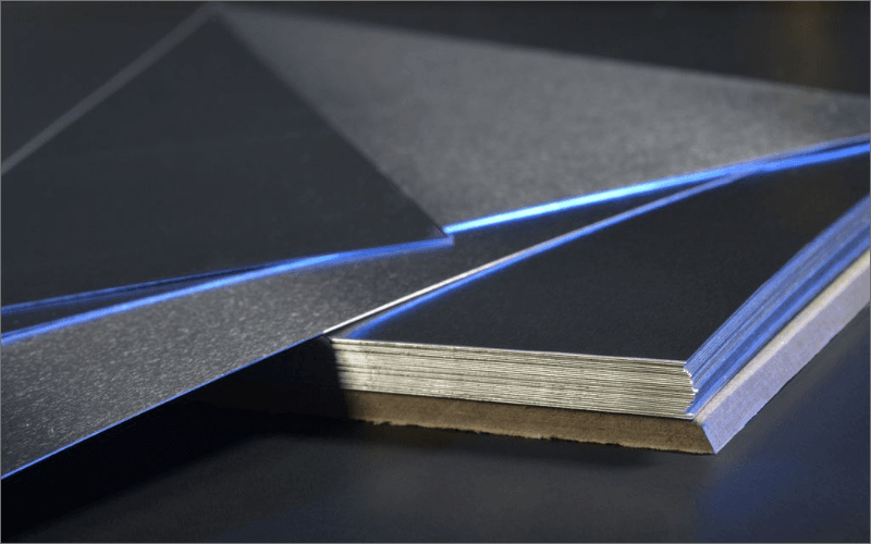 Close-up of stacked sheets of aluminum shown in various angles on a dark surface. The top of the sheets has a smooth, reflective finish, while the edges show the multiple thin layers of aluminum. A faint blue light reflects off the surface.