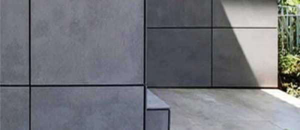 A minimalistic exterior facade of a building, featuring large square gray tiles. The sunlight casts geometric shadows across the cladding, emphasizing the clean lines and modern design. In the background, a hint of greenery is visible.