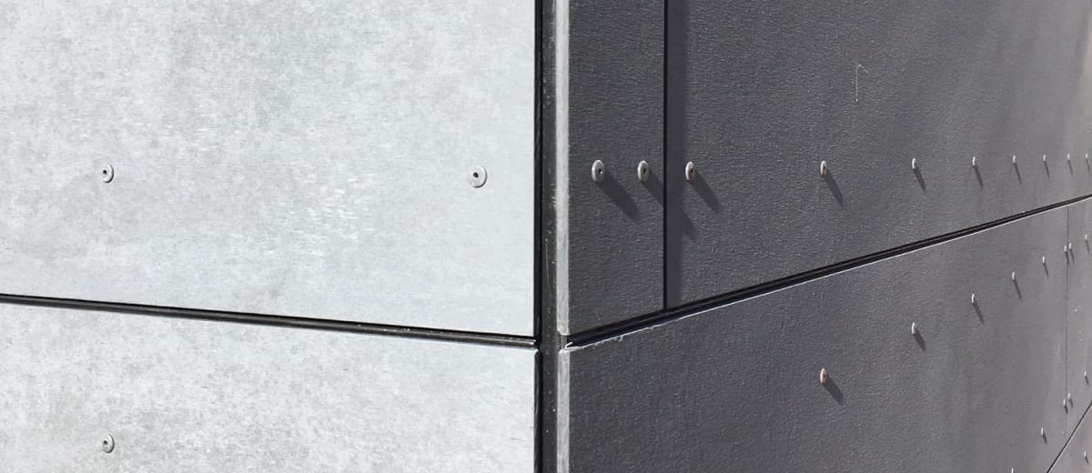 Close-up view of a building corner showcasing two different shades of grey exterior facade cladding. Each panel is fastened with visible screws, creating a grid pattern. The panels on the left are light grey while those on the right are darker grey.
