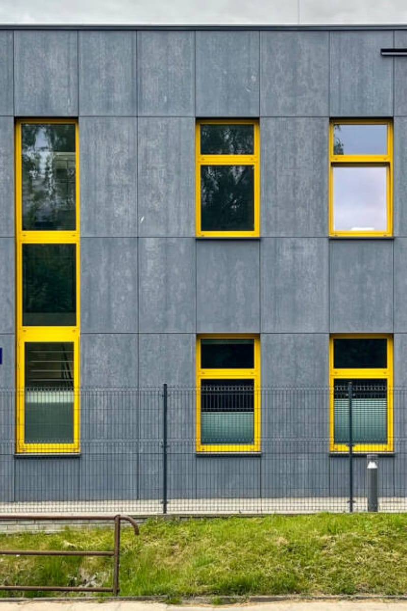 A modern, gray building with three stories, featuring tall, vertical windows framed in bright yellow. The building is surrounded by a sheet metal fence, and there is a grassy area in front. The sky is overcast, and a lone streetlight is visible near the fence.