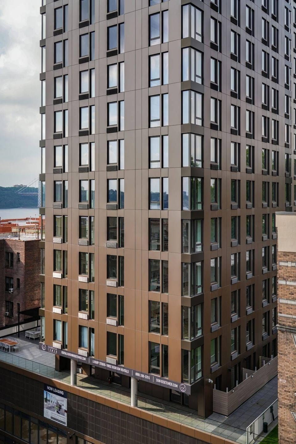 A tall, modern apartment building clad in sheet metal panels with numerous windows and a bronze-colored facade is shown. The building overlooks a body of water and adjacent structures. A “Now Leasing” sign is visible at the base, and there are potted plants on the terrace.