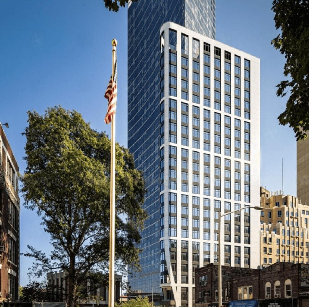 A tall, modern skyscraper with a glass facade stands prominently against a clear blue sky. In the foreground, an American flag on a pole waves gently, and trees partially frame the view. The surrounding buildings at the base add to the urban landscape's charm.