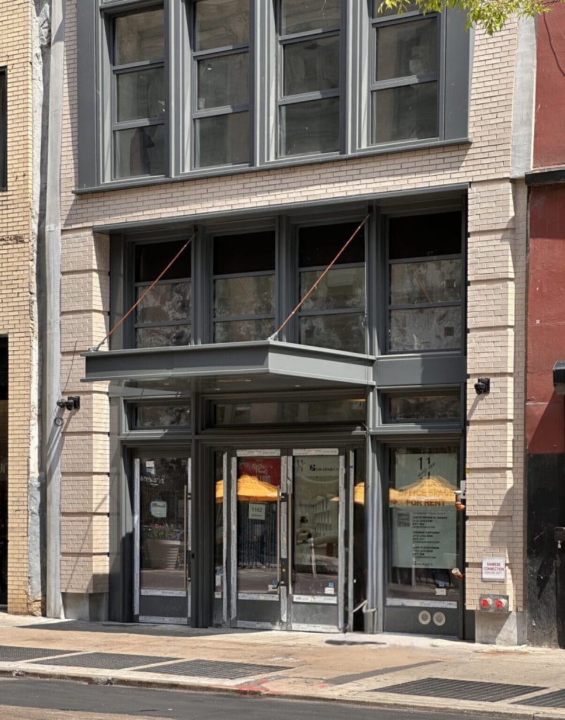 A street-level view of a building entrance with large glass windows and doors. The facade is composed of light-colored bricks, sheet metal, and dark, framed glass panels. A sign on the door indicates a rental availability. A small awning covers the entrance.