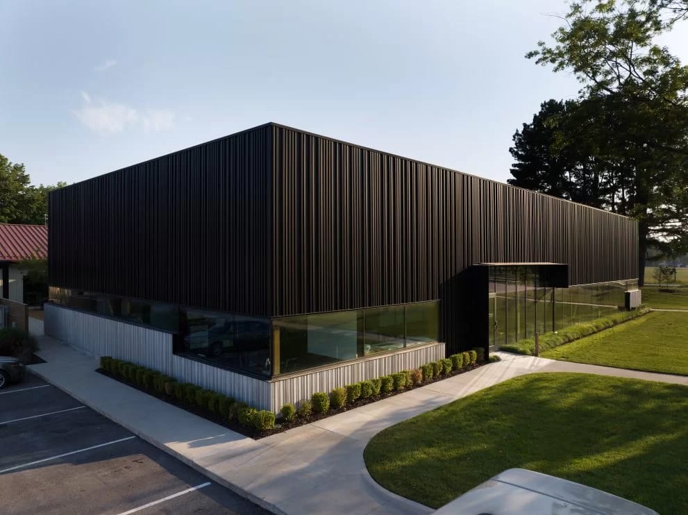 A modern, one-story commercial building with a sleek, black corrugated metal exterior facade cladding and large glass windows at the front. It is surrounded by a well-maintained lawn and small shrubs, with a paved driveway and parking area adjacent to it. Trees are visible in the background.