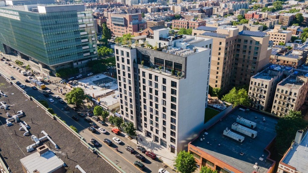 An aerial view of a cityscape featuring a tall, modern building with a rooftop terrace surrounded by other mid-rise buildings. Several cars are visible on the main road below. The building's sleek design incorporates standing seam sheet metal panels. Green spaces and mixed-use areas can be seen in the background.