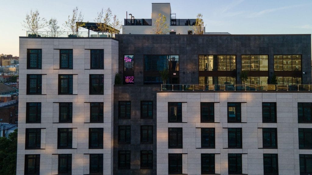 A modern multi-story building features a mix of dark and light exterior facade cladding. Numerous windows are visible, and there are trees and an outdoor seating area on the rooftop. The surrounding urban area can be partially seen in the background during sunset.