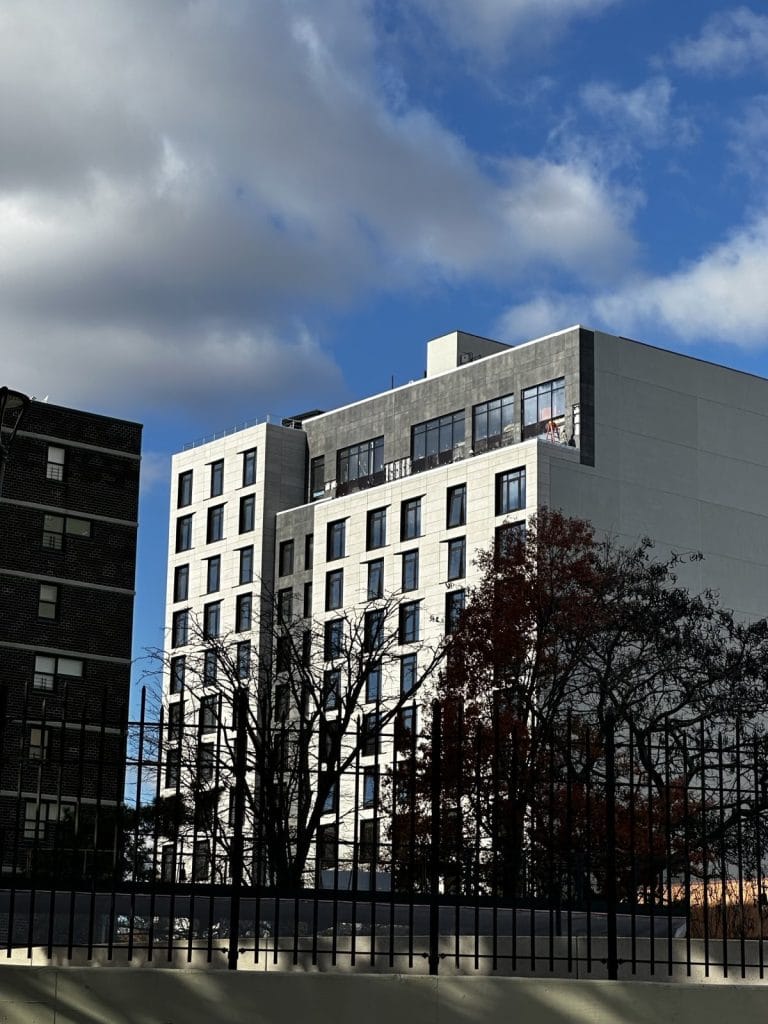 A modern, multi-story building stands tall against a partly cloudy sky. The structure features large windows and a minimalist design, incorporating standing seam elements and break metal panels. Leafless trees and a metal fence in the foreground cast shadows on a pale surface.