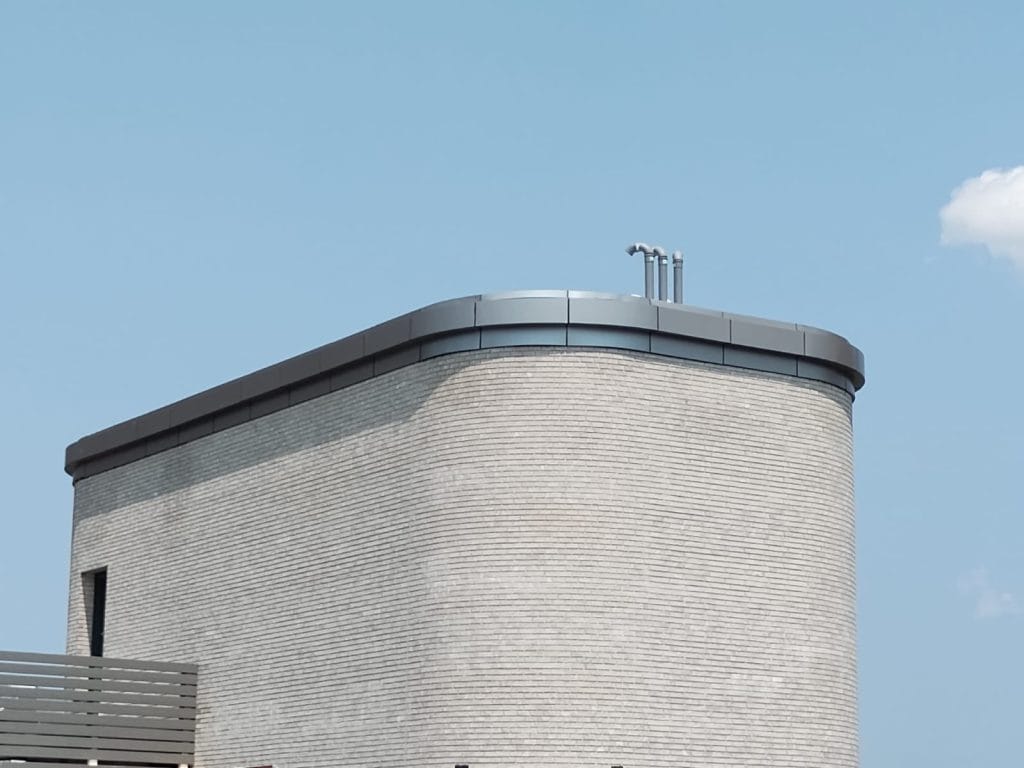 A modern, minimalist building with light gray brick walls and a rounded corner. The structure features a flat rooftop adorned with silver ventilation pipes extending upward, constructed using ACM panels. The sky in the background is clear and blue with a single white cloud.