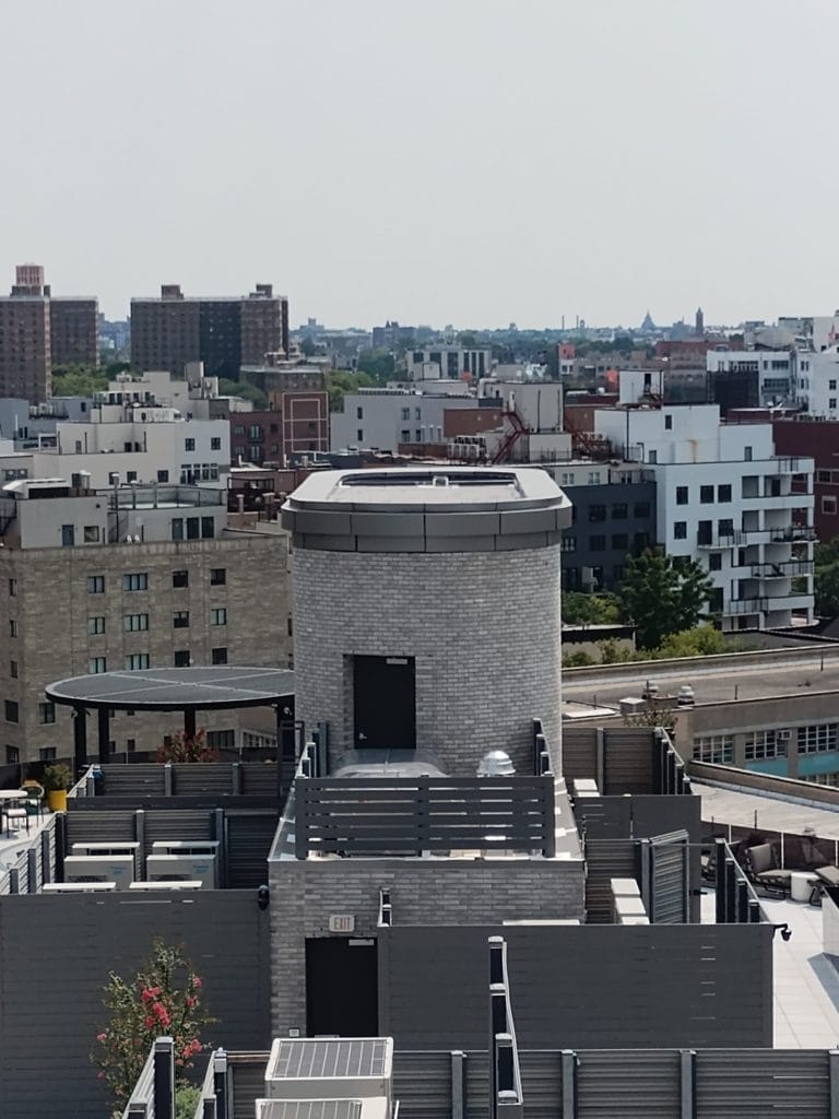 A view of a cityscape showing a variety of buildings. In the foreground, there is a cylindrical structure with sheet metal on a rooftop terrace surrounded by fencing. In the distance, numerous mid-rise and high-rise buildings are visible under a hazy sky.