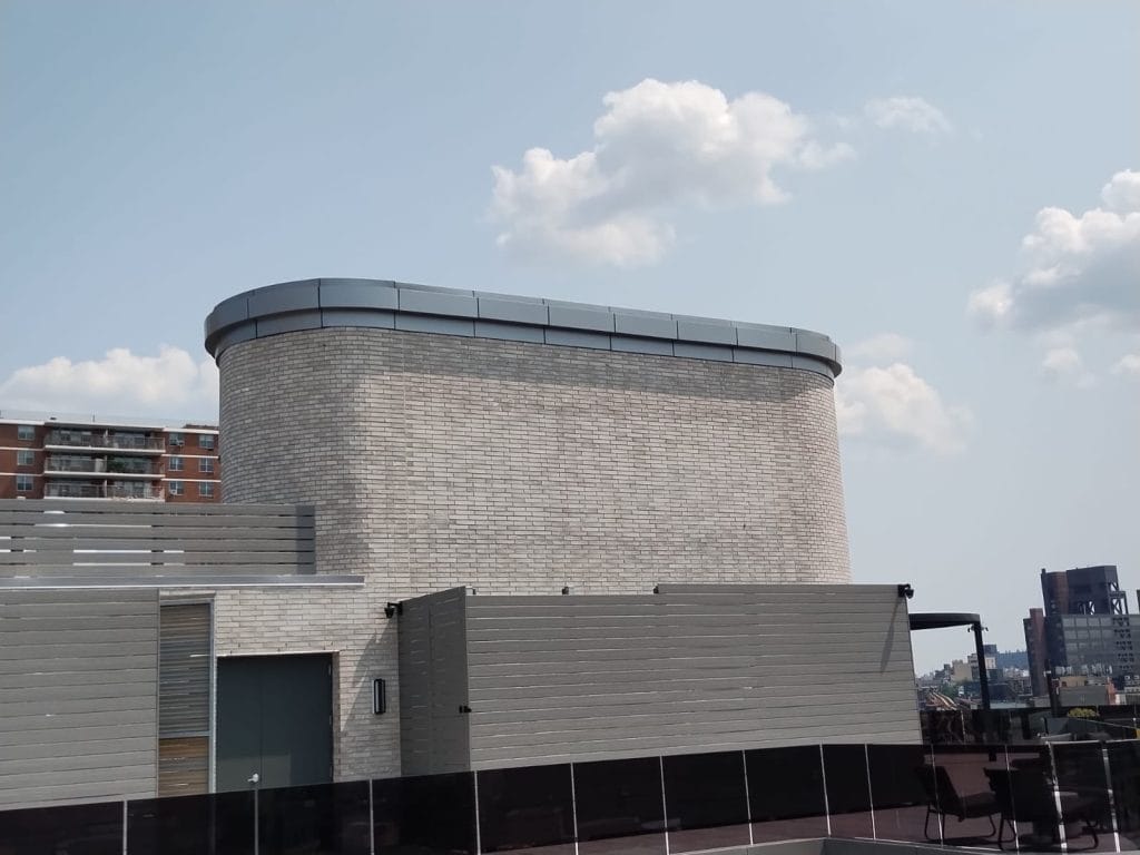 A cylindrical building with light gray brick walls and a standing seam metal roof is shown. The building is situated on a rooftop with a clear sky and scattered clouds in the background. Nearby, there are multi-story residential buildings and cityscape elements visible.