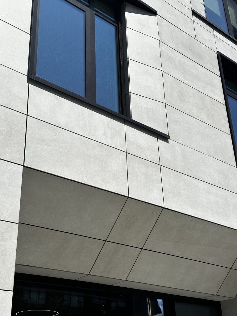 A modern building façade featuring large, light-colored rectangular tiles and dark-framed windows. The design includes an overhang above the entrance with neatly aligned tiles and sleek, minimalist architectural lines. It exudes contemporary elegance and sophistication.