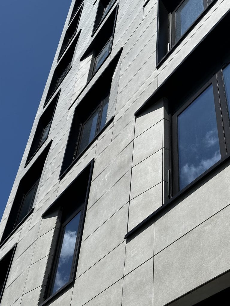 A modern, tall building with large, rectangular windows and a smooth, light-colored facade under a clear blue sky. The upward angle of the photo highlights the building's innovative design and clean lines.