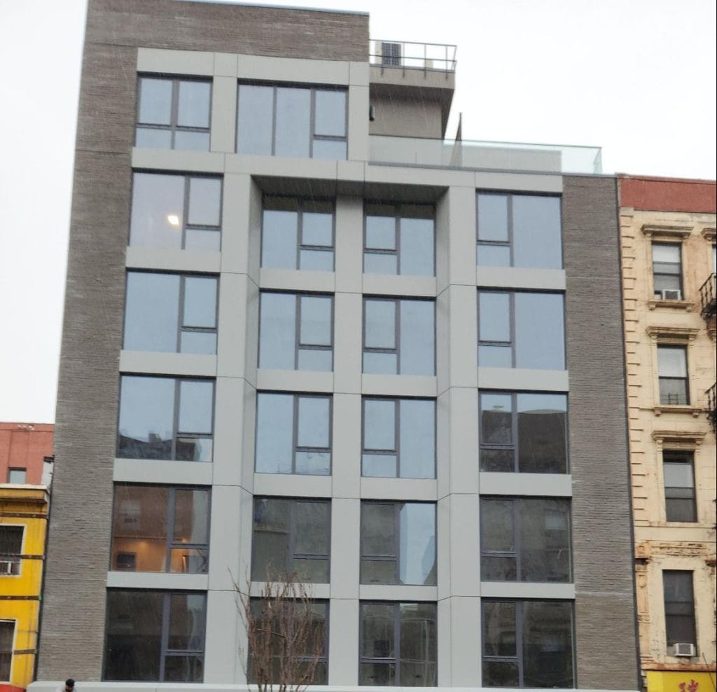 A modern multi-story building with large windows and a gray facade stands between a yellow building on the left and a beige building on the right. The structure features geometric design elements and a rooftop area perfect for events. One bare tree is visible in the foreground.