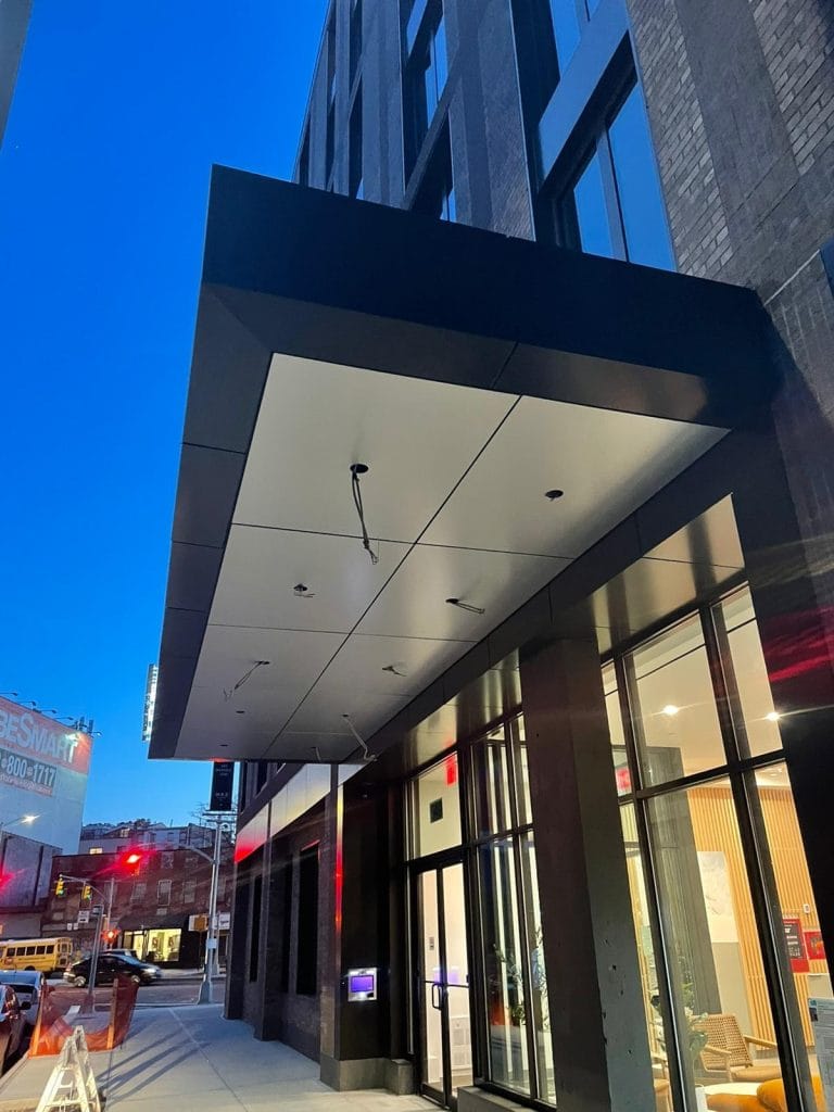 A modern building entrance at dusk with a large, flat awning extending over the sidewalk. The building features expansive glass windows, and the interior lighting creates a warm glow. In the background, a bustling street scene includes a few buildings, a prominent billboard, and traffic lights.