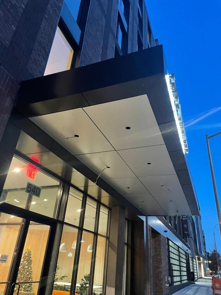 A modern building exterior at twilight features a large overhanging canopy above the entrance. The glass doors are illuminated, revealing a warmly lit interior and a decorated Christmas tree. The address "540" is subtly visible on a window near the entrance, enhancing the architectural design.