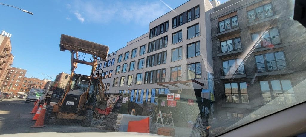 A construction site is bustling with a large excavator and safety cones. Workers are present, while the building in progress rises to five stories with completed windows. Barricades, signs, and scaffoldings ensure the area remains safe for pedestrians and traffic nearby.