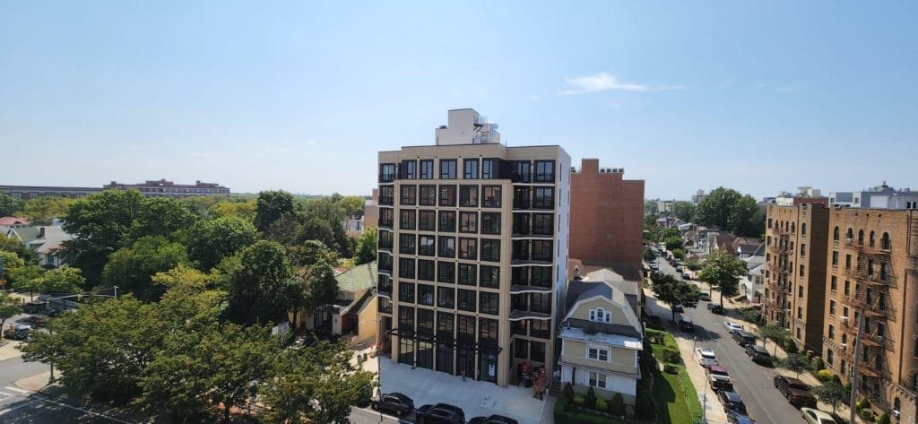 A modern, multi-story apartment building with large windows stands amidst a neighborhood of smaller homes. Trees surround the area, and the sky is clear and blue. The building boasts an elegant design and a small parking area in front, creating a perfect blend of contemporary living.