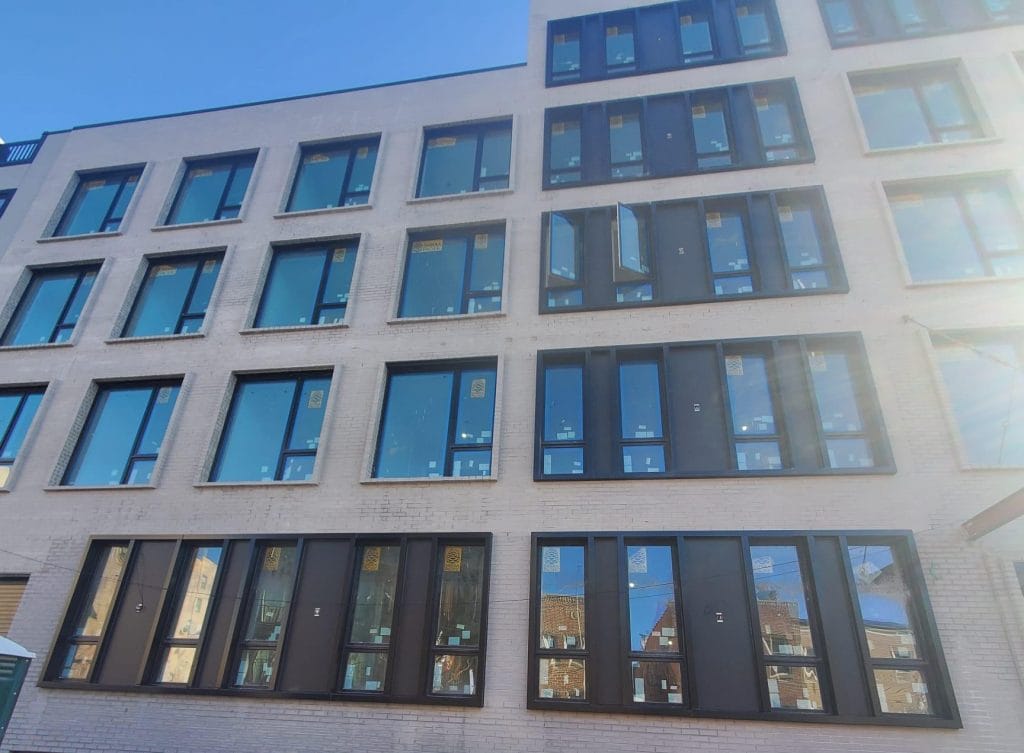 A multi-story building with numerous large windows, some of which are partially open. The facade is light-colored, and the clear blue sky is visible in the background. The windows reflect surrounding buildings and sunlight, giving it a sleek and modern appearance.