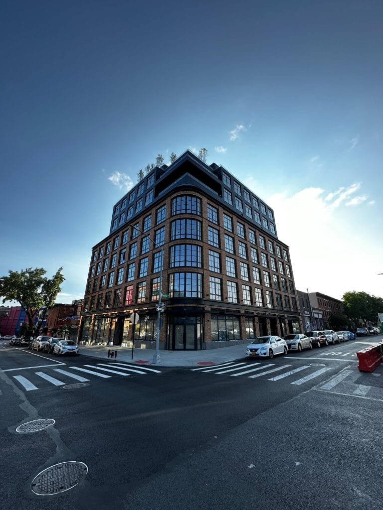 A modern, multi-story building stands at a street corner under a clear sky. The sleek structure features large windows and a mix of brick and glass facade. Cars are parked along the roadside, and trees add greenery to the left.
