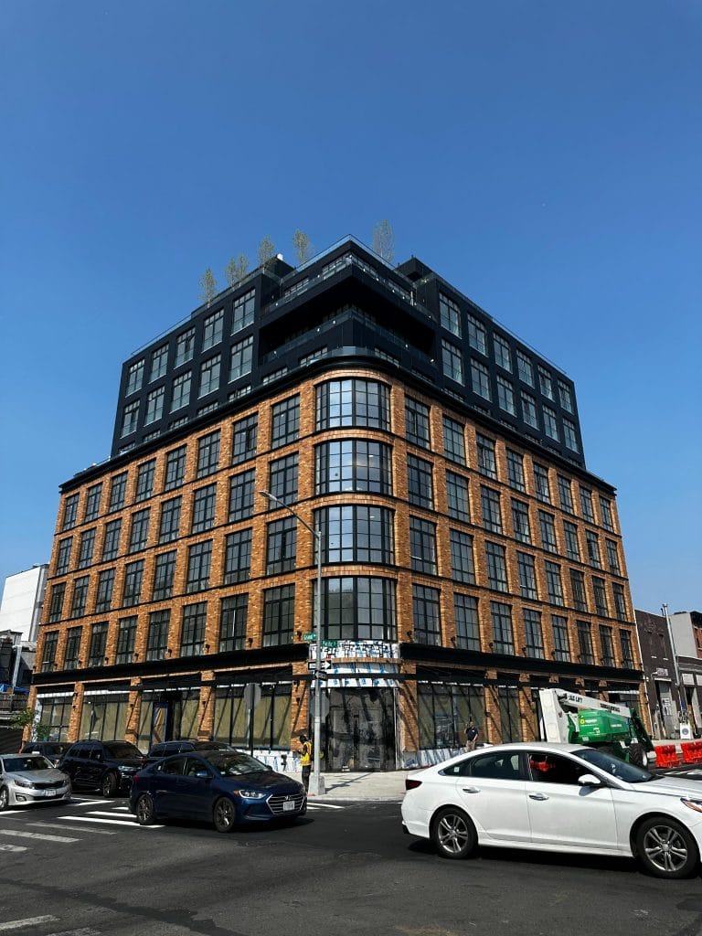 A multi-story building with a modern design features a grid of large windows and brick walls on the lower floors. The higher floors have a darker exterior. Cars are parked along the street, and a green construction vehicle is nearby. The clear blue sky enhances the urban landscape.