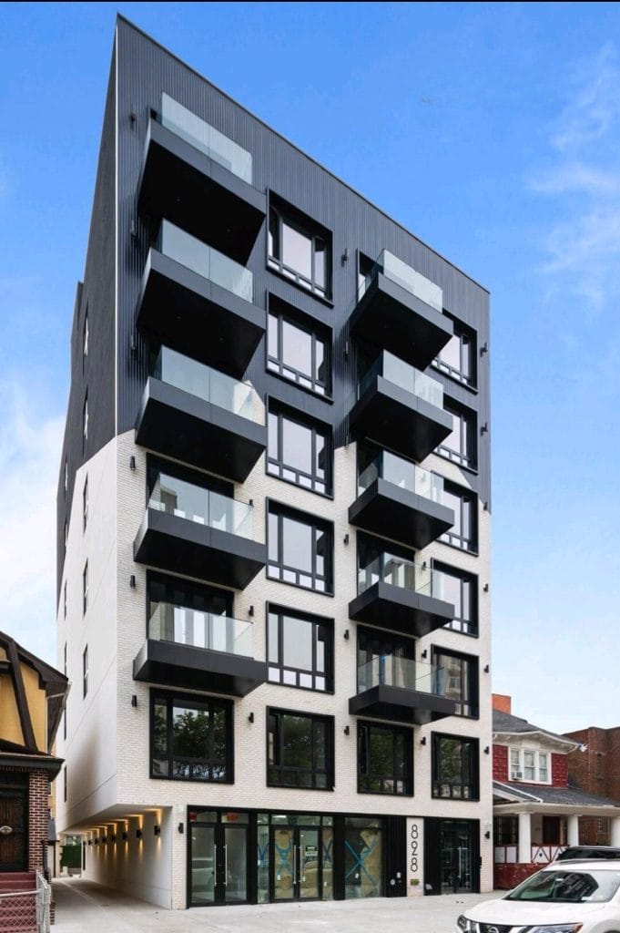 A modern multi-story apartment building with an angular design and large glass windows. The façade showcases alternating black and white sections with sleek glass balconies. The structure stands tall on a clear day against a vibrant blue sky, embodying urban elegance.