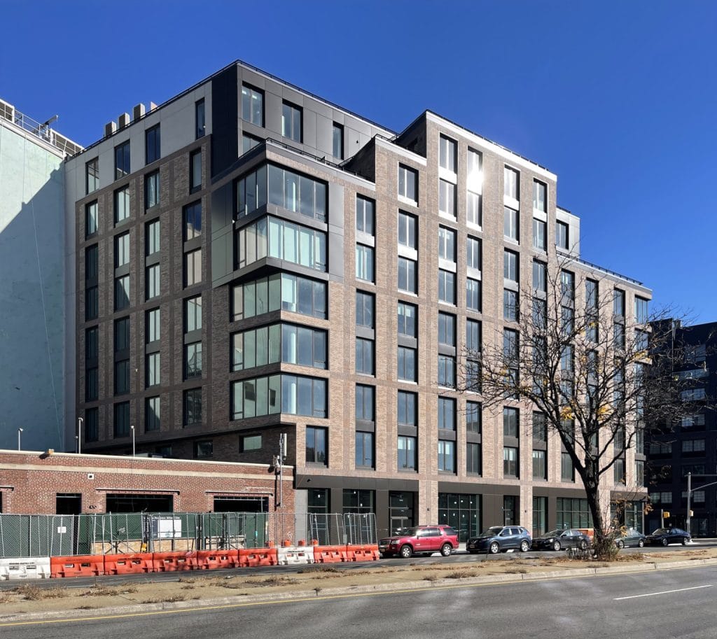 A modern multi-story apartment building with large windows and a beige-brick facade stands along a bustling city street. The clear blue sky highlights the building's sharp, contemporary design. Construction barriers and fencing in the foreground indicate ongoing development activity nearby.