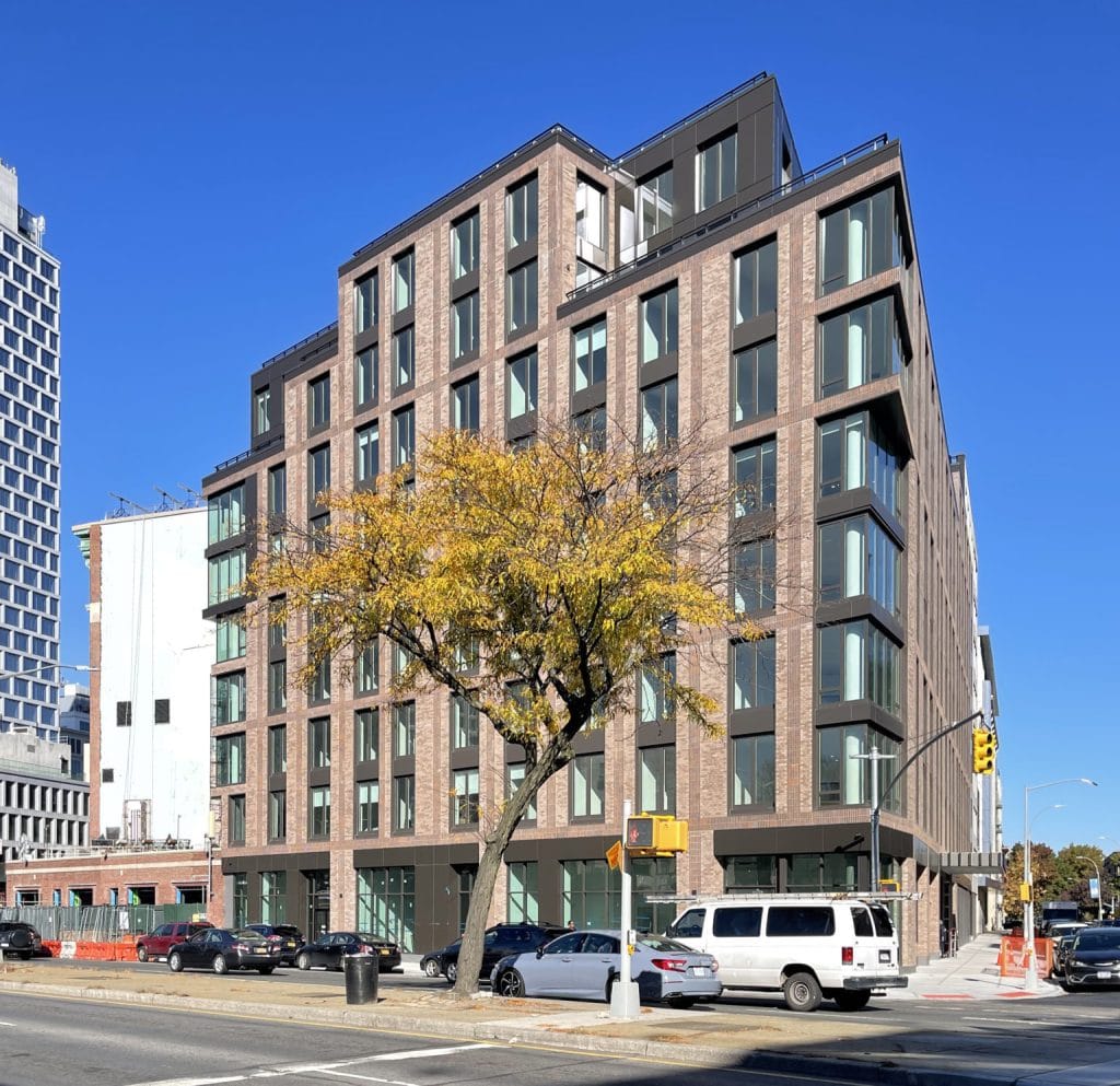 A multi-story modern apartment building with large windows and a brick facade stands on a street corner. A leafless tree is in front of the building. Several parked cars and a street light are visible under the clear, blue sky. Nearby, cyclists ride by on their morning commute.