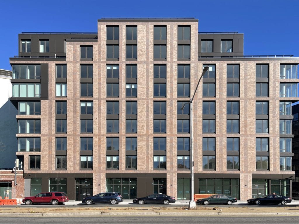 A modern multi-story apartment building with a brick facade, large windows, and a minimalist design. Several cars are parked along the street in front of the building. The sky is clear and blue, creating a serene urban atmosphere.