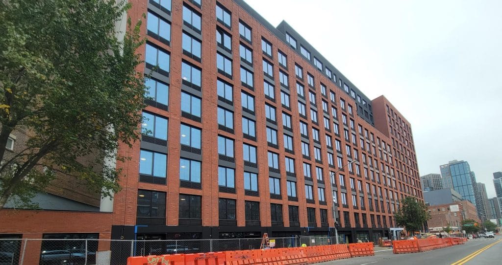 A multi-story brick apartment building with large windows stands alongside a street lined with orange construction barriers and fencing. Trees are visible on both sides of the building, while modern skyscrapers loom in the background, adding to the urban landscape.