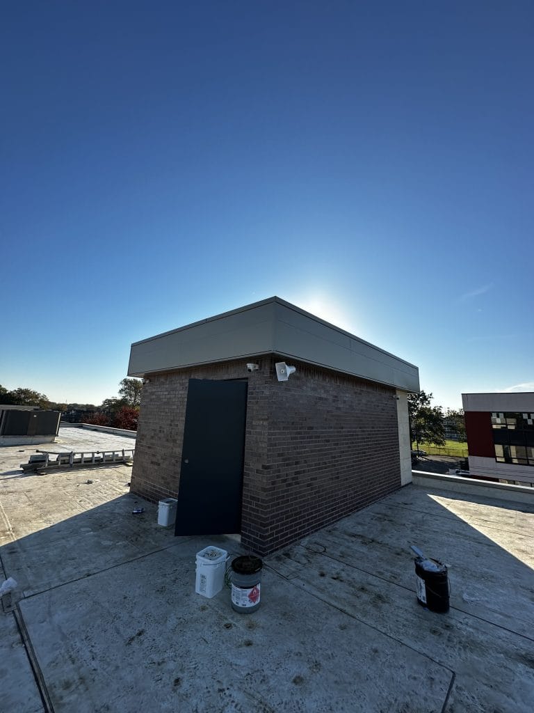 A small utility building with a brick exterior and a metal door sits on a rooftop under a clear blue sky. Sunlight illuminates the scene from behind the structure. Various maintenance supplies, like a bucket and paint can, are scattered nearby, hinting at recent touch-ups to the rooftop’s appearance.