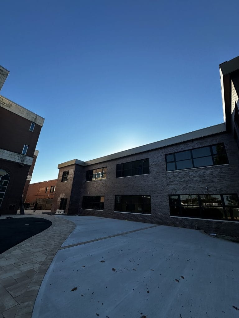 A modern, multi-story brick building with large, dark-framed windows sits under a clear blue sky. A curved pathway bordered by paved stones leads toward the structure. An additional nearby building is visible to the left, partially shadowed by trees.