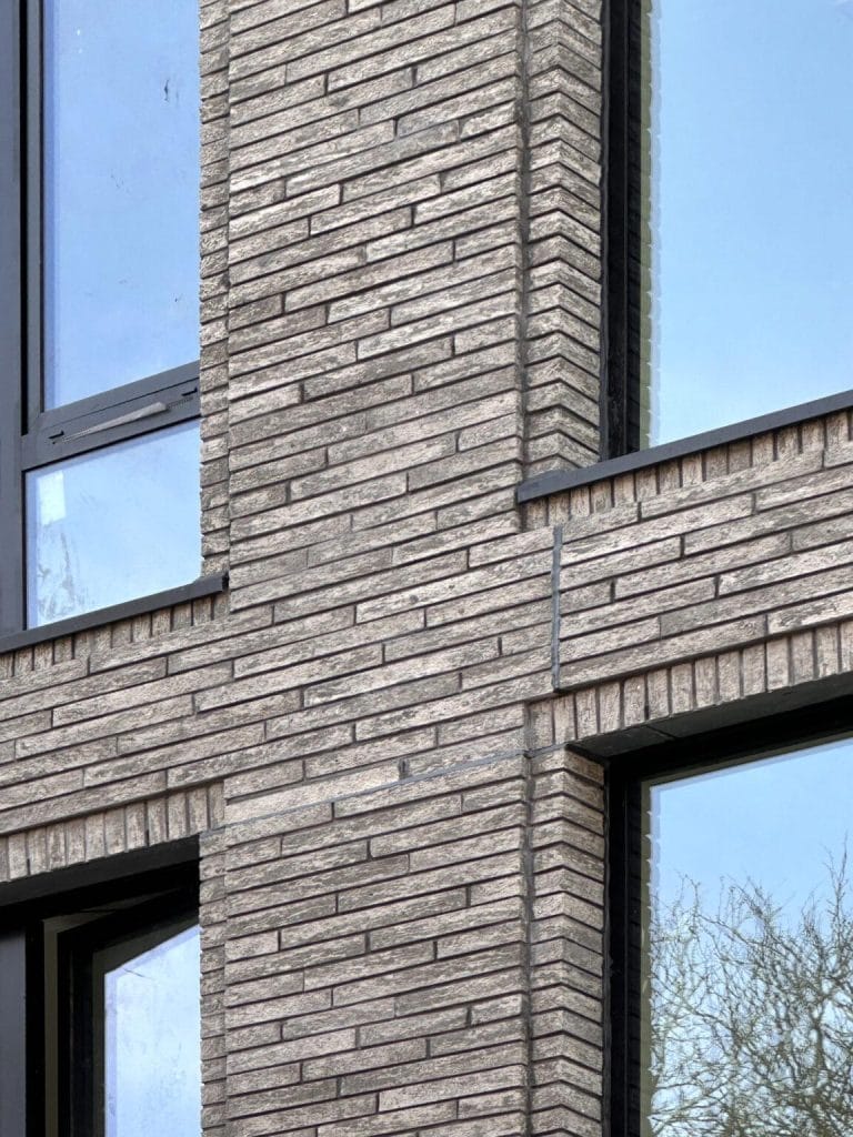 A close-up view of a modern building's facade, showcasing a mix of vertical and horizontal narrow brick patterns. The building features large, reflective glass windows accented with standing seam sheet metal. The scene captures clear blue skies and some tree branches reflected in the windows.