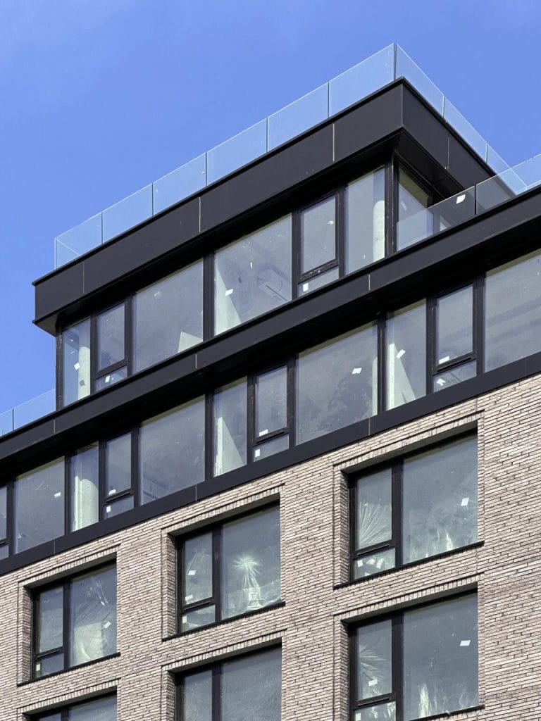 A multi-story modern building with large glass windows and a beige brick facade. The top floor, adorned with floor-to-ceiling glass panels and a terrace featuring a glass railing, also incorporates sleek sheet metal accents. The sky in the background is clear and blue.