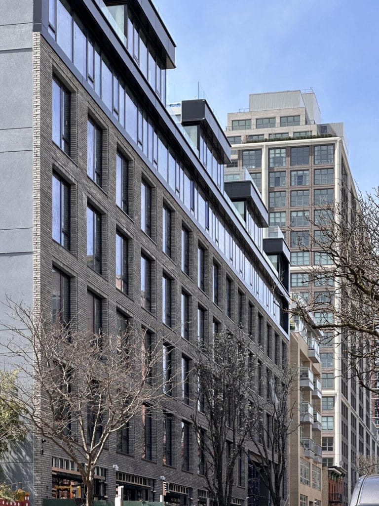 A city street with modern, multi-story buildings. The foreground features a dark gray brick building with large windows, while a taller, lighter-colored building made of sheet metal stands in the background. Leafless trees line the street, suggesting a winter or early spring setting.
