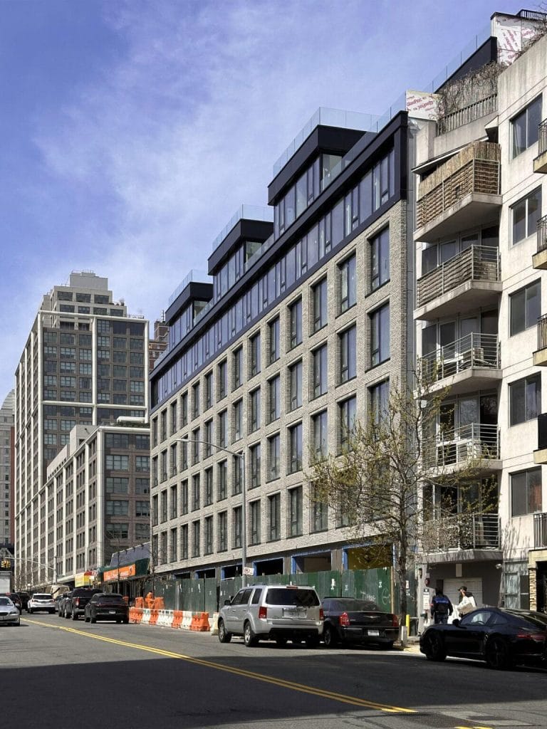 A city street features modern multi-story buildings with large windows and fiber cement accents. One building has several staggered balconies. Cars are parked along the road, and there are orange construction barriers on the sidewalk. The sky is clear and blue.