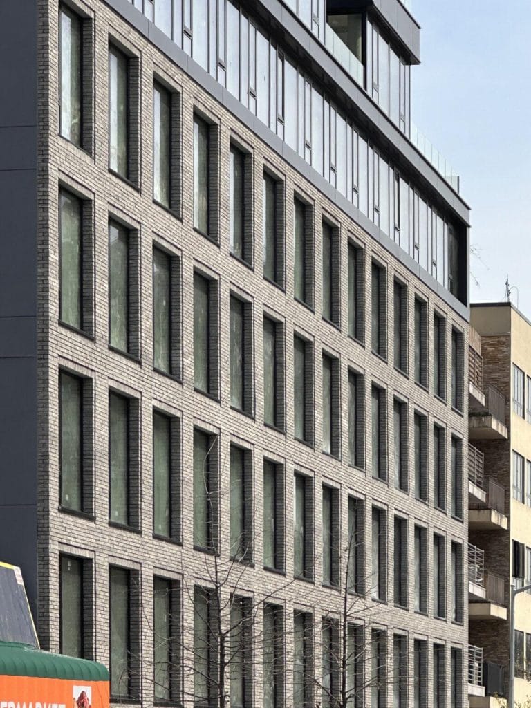 A modern apartment building with a facade of rectangular windows and a brick exterior. The structure has five floors, with the top floor featuring larger windows and possibly balconies. Nearby, a partial view of another building with balconies is visible. The exterior facade cladding includes elements like ACM panels.