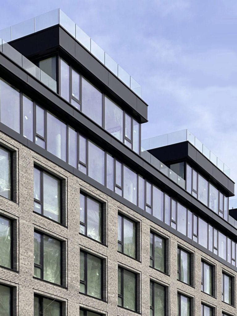 A modern multi-story building with large windows and a brick exterior. The upper floors feature floor-to-ceiling windows and rooftop terraces, while the lower floors have a uniform arrangement of rectangular windows. The clear sky above complements the sleek Exterior Facade Cladding elements.