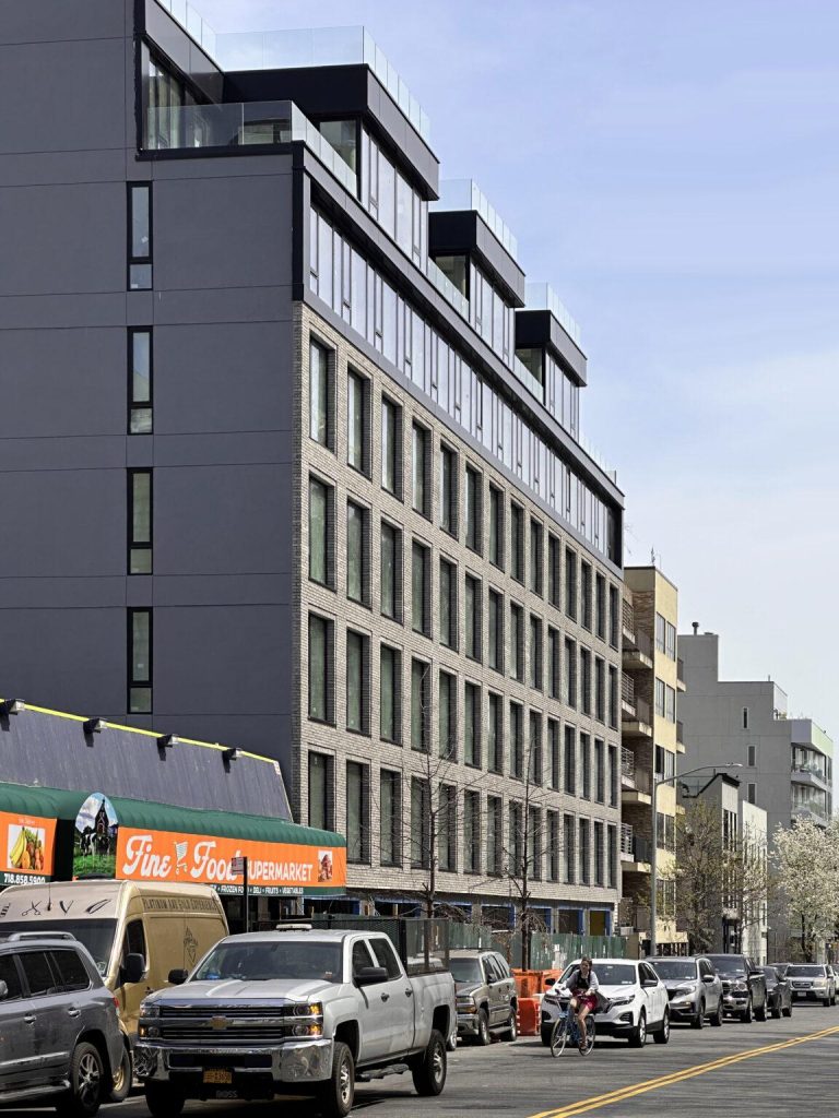 A modern, multi-story building with large windows and a mix of dark and light grey fiber cement panels stands beside a street lined with parked cars. A supermarket with an orange sign reading "Fine Fare Supermarket" is located at the ground level of the building.
