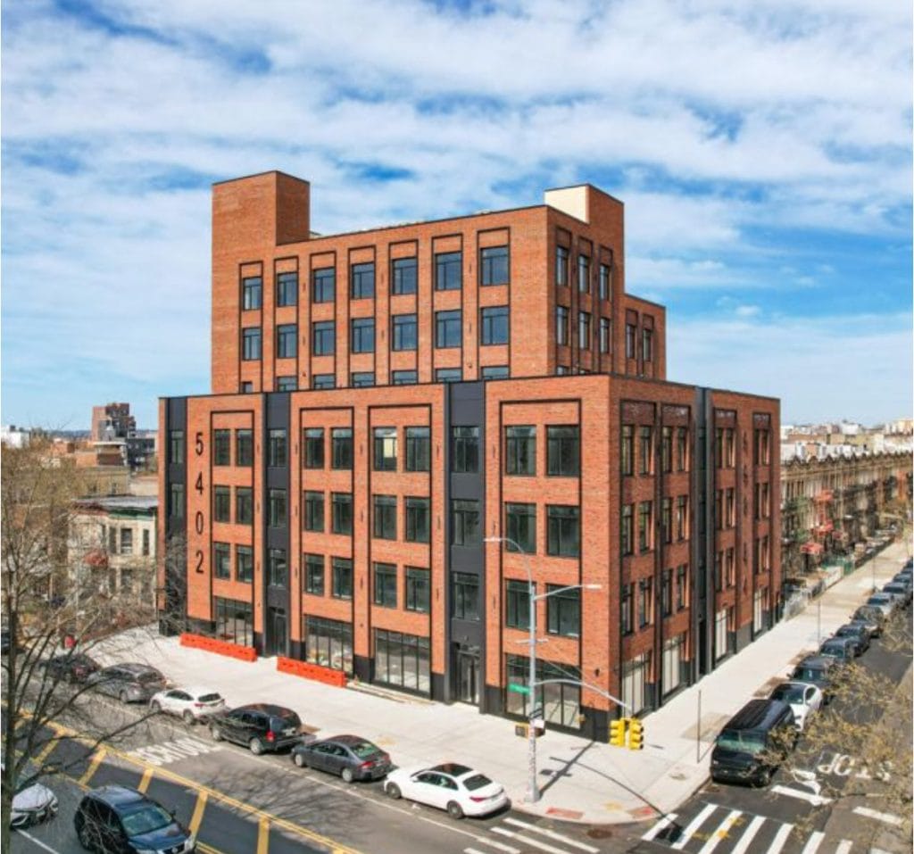 A six-story red brick building with black-framed windows sits on the corner of a city street. The building has the number "5402" displayed vertically on its fiber cement facade. Several cars are parked along the street, and a crosswalk is visible in the foreground.