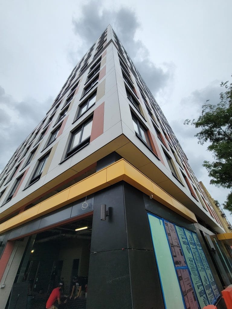 A tall, modern building with a sharp corner extends towards the sky, showcasing multiple floors with a mix of gray, white, and red Exterior Facade Cladding panels. The ground floor appears to be under construction. The overcast sky and nearby trees are visible in the background.