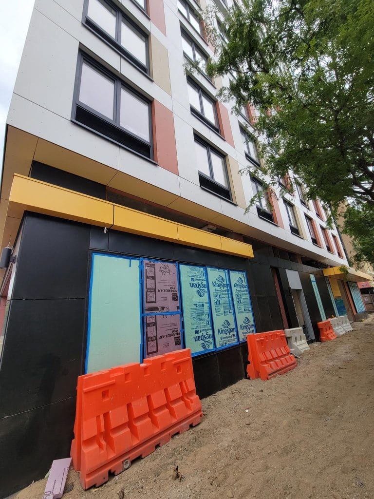 A multi-story building under construction, featuring large windows and partially finished Exterior Facade Cladding. Bright orange barricades line the sandy pathway in front, with construction material visible around the lower portion of the structure.