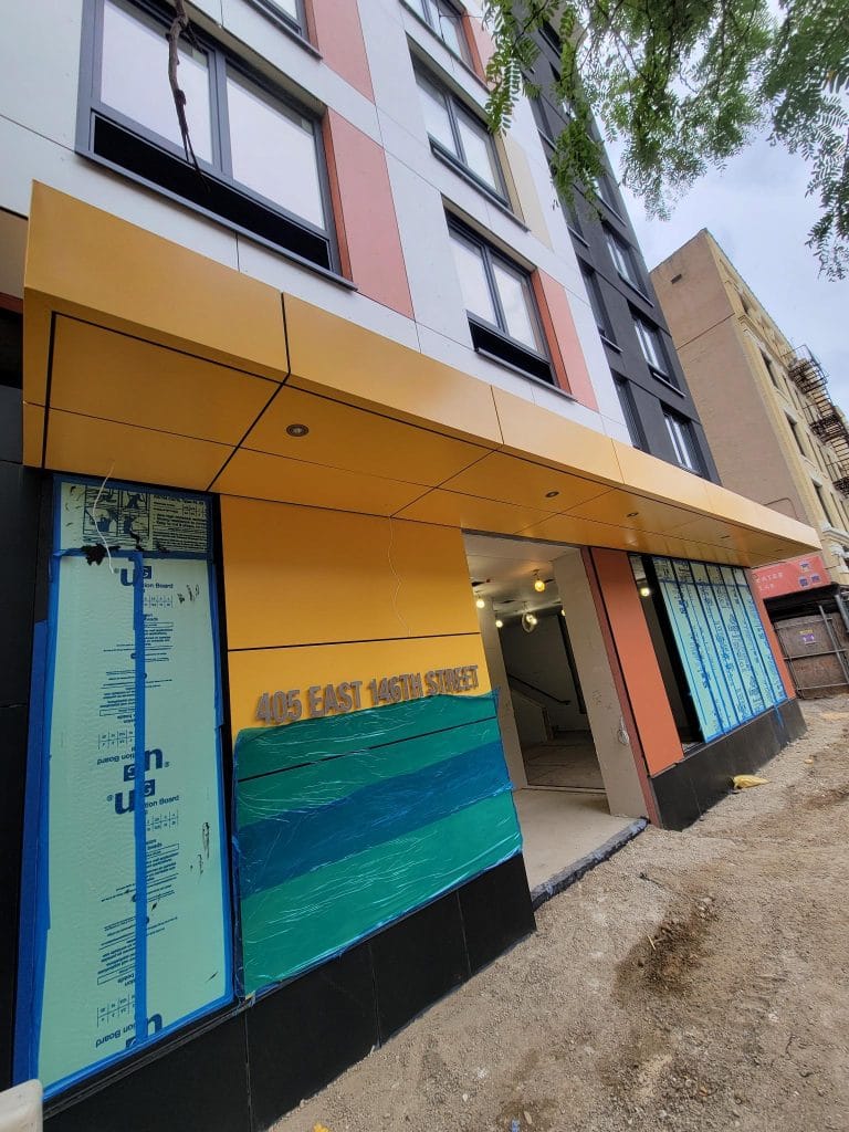 A multi-story building under construction with a partially completed entrance canopy features reflective blue and green panels made of break metal. The address "405 EAST 146TH STREET" is prominently displayed. Construction materials and scaffolding are scattered around the site.