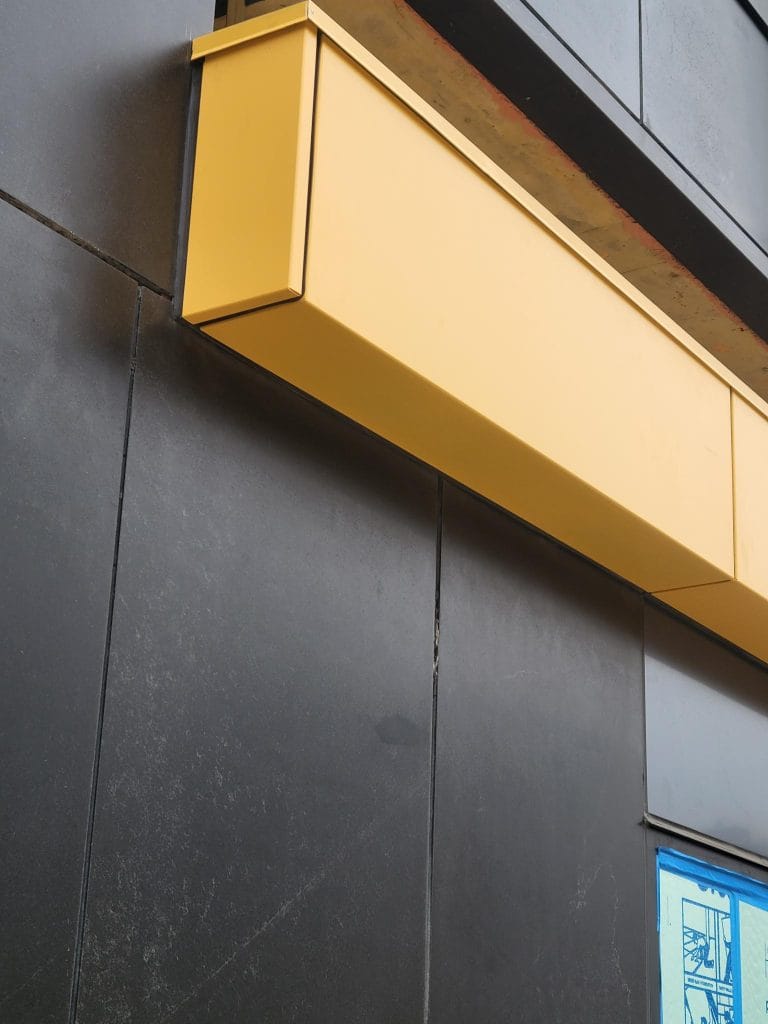 A close-up view of a modern building facade featuring a sleek, black exterior cladding. A large, rectangular, golden-yellow sign extends horizontally across the facade. At the bottom right, a small section of a blue and white illustration is visible.
