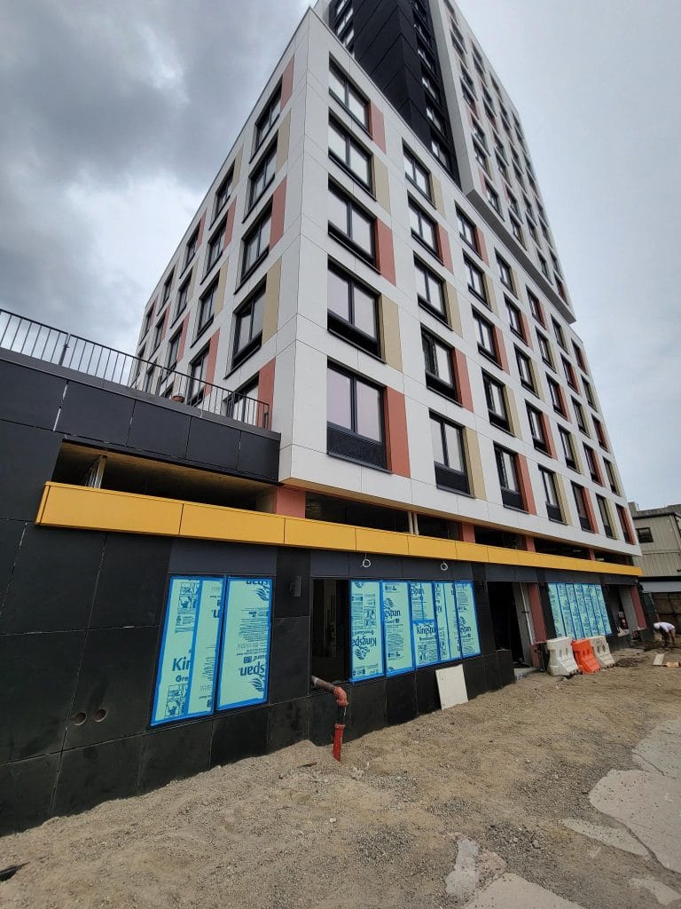 A multistory building under construction, featuring a modern design with rectangular windows and colorful panels. The ground floor windows are covered with protective film. The area around the building is unpaved, scattered with construction materials and sheet metal for the exterior facade cladding.