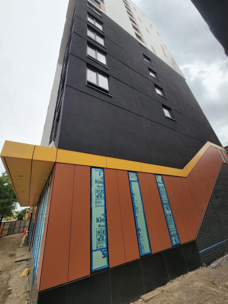 A tall, modern building under construction is seen from a low angle, emphasizing its height. The lower part features brown panels with blue insulation visible alongside sheet metal cladding, while the upper sections are painted black and white. The sky is overcast, and the ground is unpaved.