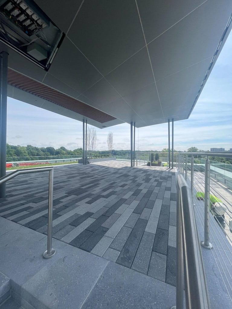 A spacious outdoor terrace with modern design elements, featuring a patterned paved floor and surrounded by sheet metal railing. The terrace is covered with a sleek standing seam roof and offers a scenic view of green trees and a distant landscape under a cloudy sky.