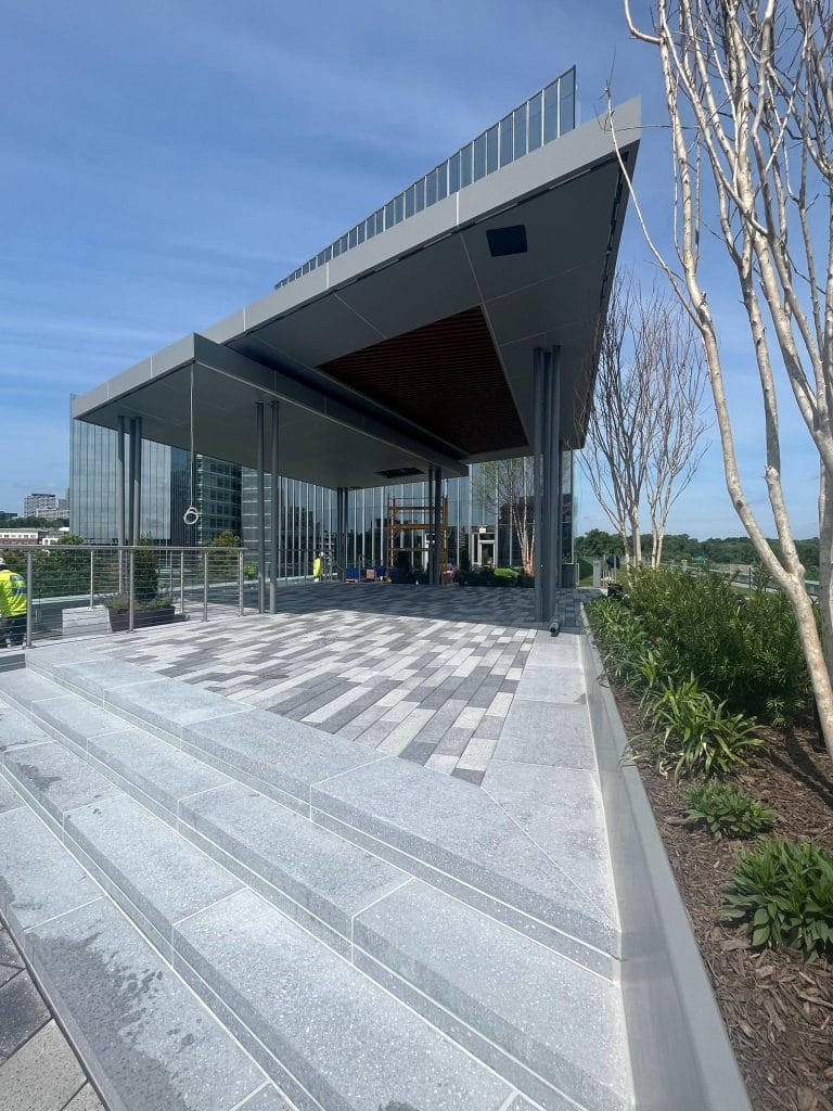 A modern building with an angular roof and large glass windows, featuring exterior facade cladding of ACM. It is situated atop a tiered platform with light gray steps. The outdoor area showcases potted plants along the side and a clear blue sky in the background.